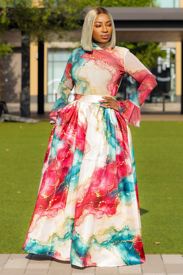 A woman wearing a long-sleeved maxi dress with a colorful abstract watercolor print in pink, teal, and cream, standing outdoors on a paved walkway with a green lawn and modern building in the background.