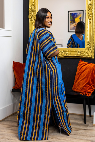 A woman wearing a long blue and gold striped kaftan dress, standing in a room with a large gold-framed mirror, orange pillows, and a wooden floor.