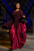 A woman wearing a floor-length burgundy gown with a sheer black lace bodice and flared sleeves, standing on outdoor stone steps at night under blue architectural lighting.