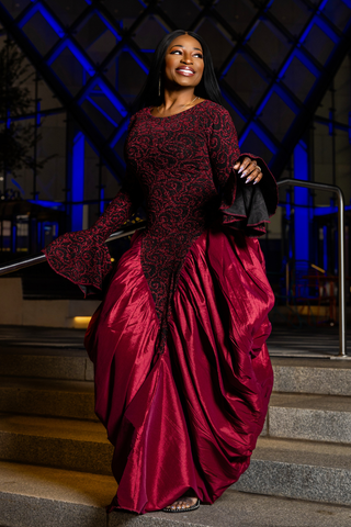 A woman wearing a floor-length burgundy gown with a sheer black lace bodice and flared sleeves, standing on outdoor stone steps at night under blue architectural lighting.