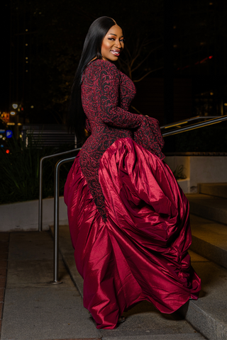 A woman wearing a floor-length formal gown with a sheer black lace bodice and a voluminous deep red satin skirt, posing on outdoor steps at night.