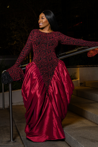 A woman wearing a long-sleeve formal gown with a black lace bodice and a voluminous deep red satin skirt, posing on outdoor stone steps at night.