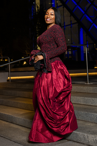 A woman posing on outdoor stone steps at night, wearing a floor-length formal gown with a dark red and black patterned lace bodice and a voluminous deep red satin skirt.