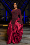 A woman wearing a floor-length formal gown with a black lace bodice and a voluminous deep red satin skirt, standing on stone steps at night with blue-lit architectural background.