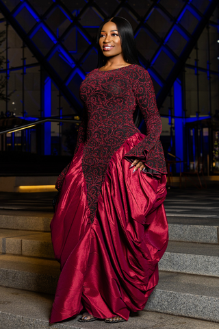 A woman wearing a floor-length formal gown with a black lace bodice and a voluminous deep red satin skirt, standing on stone steps at night with blue-lit architectural background.