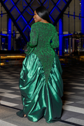 A woman wearing a voluminous emerald green evening gown with a sheer lace bodice and a shiny satin skirt, photographed from behind in an indoor venue with blue lighting.