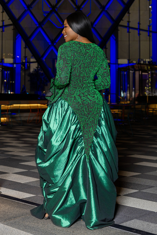 A woman wearing a voluminous emerald green evening gown with a sheer lace bodice and a shiny satin skirt, photographed from behind in an indoor venue with blue lighting.