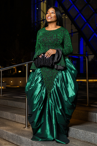 A woman wearing a formal emerald green gown with a lace bodice and a voluminous satin skirt, holding a black clutch on outdoor stone steps at night.