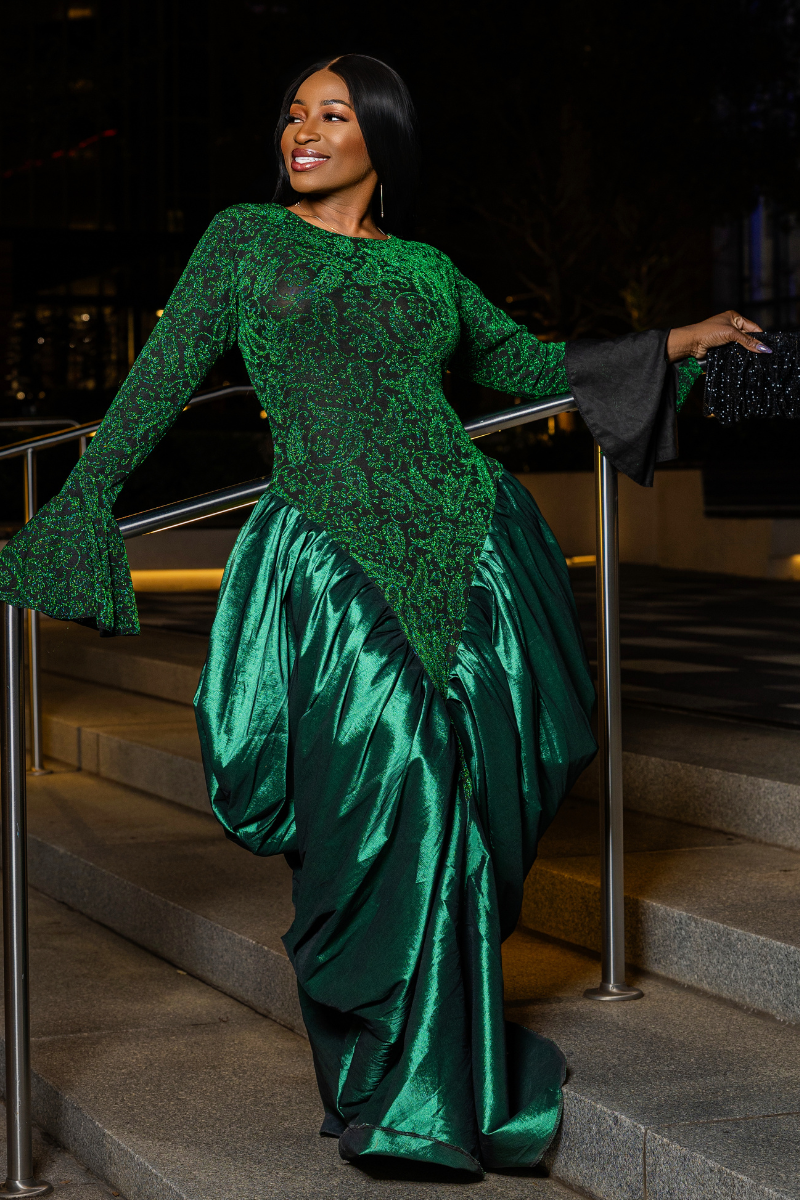 A woman posing on outdoor stone steps at night wearing a floor-length emerald green gown with a sheer lace long-sleeve bodice and a voluminous satin skirt.