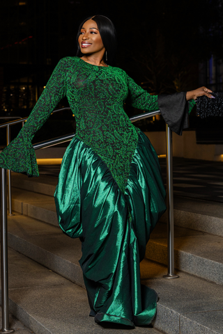 A woman posing on outdoor stone steps at night wearing a floor-length emerald green gown with a sheer lace long-sleeve bodice and a voluminous satin skirt.