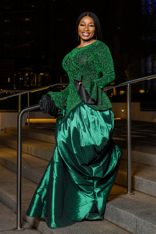 A woman stands on outdoor stone steps at night wearing a voluminous emerald green dress with a textured lace long-sleeve top and black gloves.