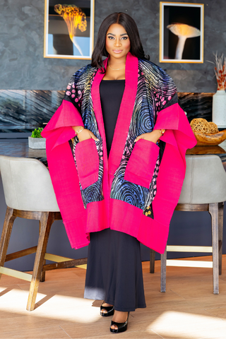 A woman modeling a vibrant hot pink open-front cardigan with contrasting black, blue, and white patterned panels and large front pockets, styled over a black dress in a modern indoor setting.