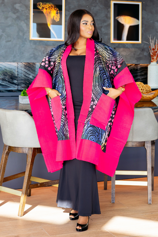 A woman models a vibrant hot pink and black patterned open-front cardigan or wrap in a modern indoor setting with gray chairs and framed wall art.