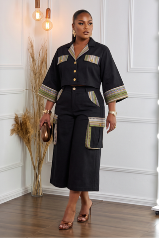 A woman models a black two-piece outfit consisting of a short-sleeve button-front top and matching wide-leg pants, both featuring beige and olive green patterned trim details. She is standing in a room with white paneled walls, wooden floors, and decorative dried grass in a vase.