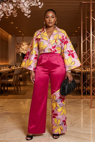 Woman in a colorful floral blouse and pink pants standing in an elegant interior setting.