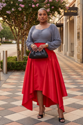 Woman in a red skirt and gray top standing on a sidewalk with a store sign in the background.