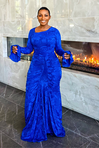 A woman wearing a royal blue long-sleeve maxi dress with a textured jacquard pattern, standing in front of a modern fireplace with marble walls.
