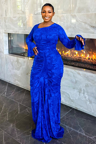 A woman wearing a royal blue long-sleeve maxi dress with ruching stands in front of a modern fireplace with marble walls.