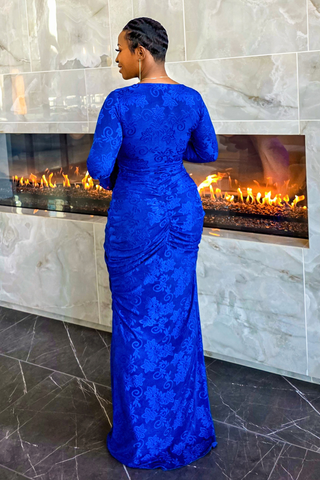 A woman stands with her back to the camera wearing a royal blue lace long-sleeve floor-length gown in front of a modern fireplace with marble walls.