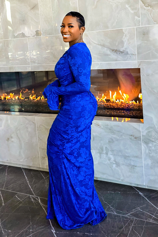 A woman wearing a royal blue long-sleeve lace maxi dress, standing in front of a modern fireplace with a marble tile wall.