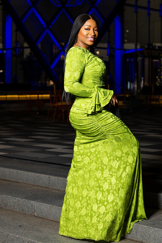 A woman wearing a bright lime green long-sleeve maxi dress with a textured floral jacquard pattern, standing on outdoor stone steps at night with a modern glass building and blue lighting in the background.