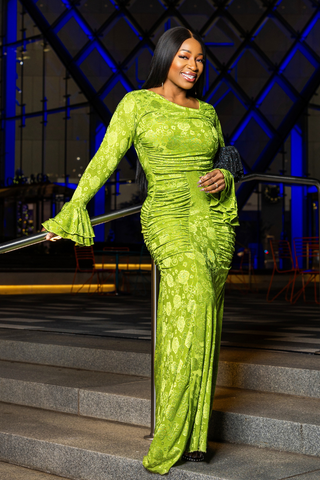 A woman wearing a bright lime green long-sleeve maxi dress with a floral lace overlay stands on outdoor stone steps at night, holding a black handbag.
