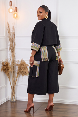 A woman wearing a black two-piece outfit with colorful patterned trim and fringe details, standing in a decorated room with a white paneled wall, wooden floor, and dried grass decor.