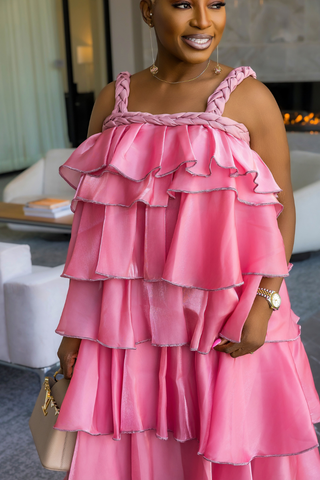 A woman wearing a bright pink tiered ruffle dress with braided shoulder straps, holding a beige handbag in a modern indoor setting.