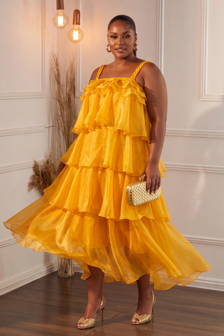A woman modeling a bright yellow tiered tulle dress with thin shoulder straps, holding a small gold clutch and wearing gold heels in an indoor room with white paneled walls and warm hanging lights.