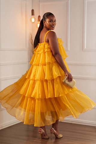 A woman wearing a bright yellow tiered ruffle dress, holding a small gold clutch, standing in a room with white paneled walls and wooden floors.