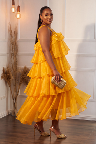 A woman modeling a bright yellow tiered ruffle maxi dress with a halter neckline, holding a small woven clutch and wearing gold heels in an indoor setting with a white paneled wall.