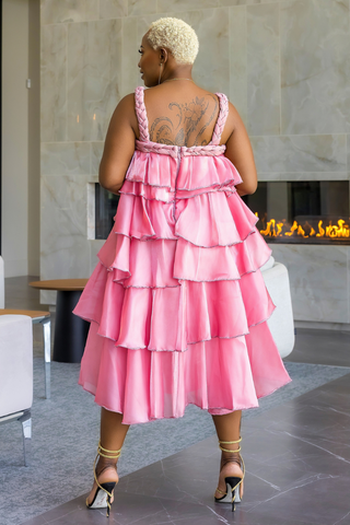 A woman viewed from behind wearing a bright pink tiered ruffle dress with braided straps, standing in a modern indoor setting with a fireplace and marble walls.