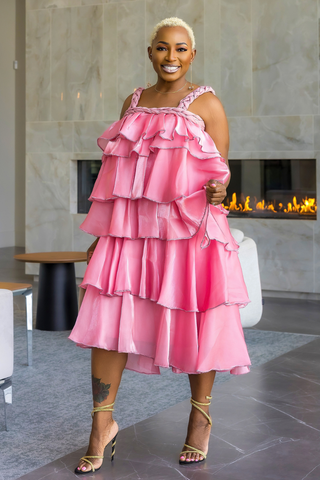 A woman wearing a bright pink tiered ruffle dress with a low-rise hem and gold strappy heels, standing in a modern indoor setting with a fireplace in the background.