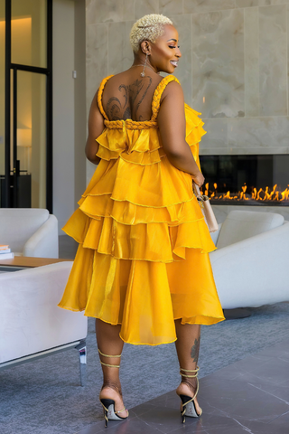 A woman wearing a bright yellow tiered ruffle dress, photographed from behind in a modern living room setting with a fireplace and white sofas.