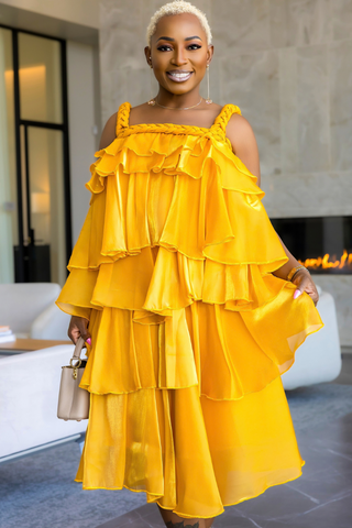 A woman wearing a bright yellow tiered ruffle dress with cold-shoulder straps, holding a small beige handbag in a modern indoor setting.