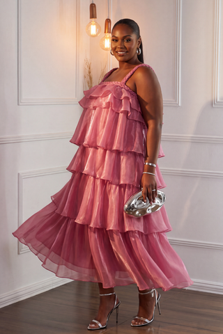 A woman modeling a pink tiered ruffle midi dress with a smocked bodice, paired with metallic silver high heels and a matching metallic clutch, standing in a white paneled room.
