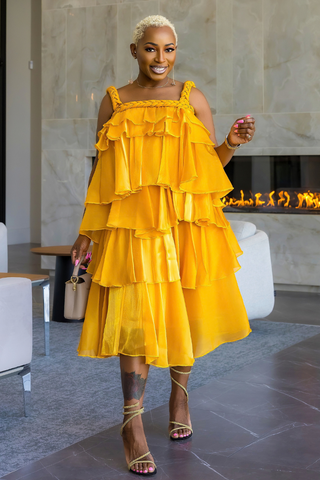 A woman wearing a bright yellow tiered ruffle dress with cold-shoulder straps, paired with tan lace-up heeled sandals, standing in a modern indoor setting with a fireplace and marble walls.