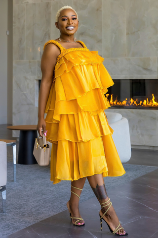 A woman models a bright yellow tiered ruffle dress in a modern indoor setting with a fireplace in the background.