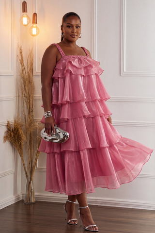 A woman models a pink tiered ruffle midi dress with spaghetti straps, paired with silver high-heeled sandals and a silver clutch, standing in a white paneled room with decorative dried grass in a vase.