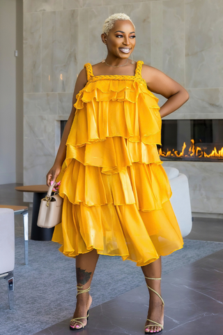 A woman models a bright yellow tiered ruffle dress in a modern indoor setting with a fireplace in the background.