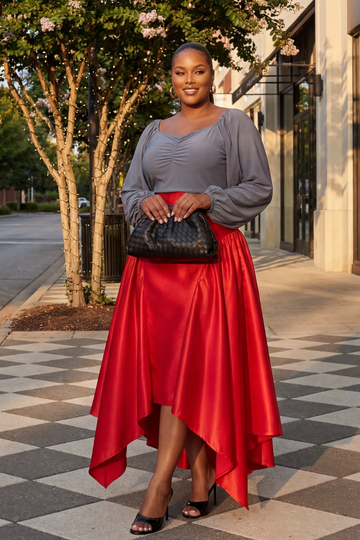 Woman in a gray top and red skirt standing on a sidewalk with trees and buildings in the background.