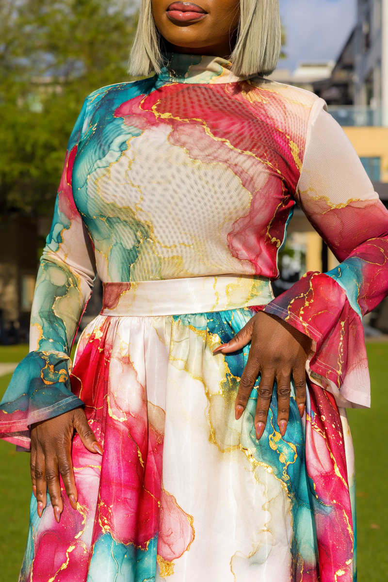 A woman wearing a long-sleeve high-neck dress with a colorful abstract marble print in pink, teal, cream, and gold, photographed outdoors on a grassy lawn.