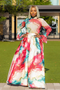 A woman stands outdoors on a paved walkway in front of a grassy lawn and building, wearing a long-sleeved maxi dress with a colorful abstract watercolor print in pink, teal, blue, and white, featuring a white waist belt.