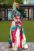 A woman stands outdoors on a paved walkway in front of a grassy lawn and modern building, wearing a long-sleeved high-neck maxi dress with a colorful abstract watercolor print in teal, pink, red, and cream.