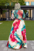 A woman viewed from behind wearing a long-sleeved maxi dress with a vibrant abstract watercolor print in pink, teal, red, and gold on a cream base, standing on a paved walkway in front of a modern outdoor setting.