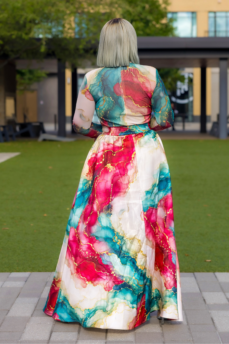 A woman viewed from behind wearing a long-sleeved maxi dress with a vibrant abstract watercolor print in pink, teal, red, and gold on a cream base, standing on a paved walkway in front of a modern outdoor setting.