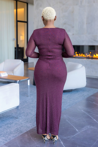 A woman viewed from behind wearing a long-sleeve, floor-length burgundy dress with a textured, dotted pattern, standing in a modern living room with a fireplace and white chairs.