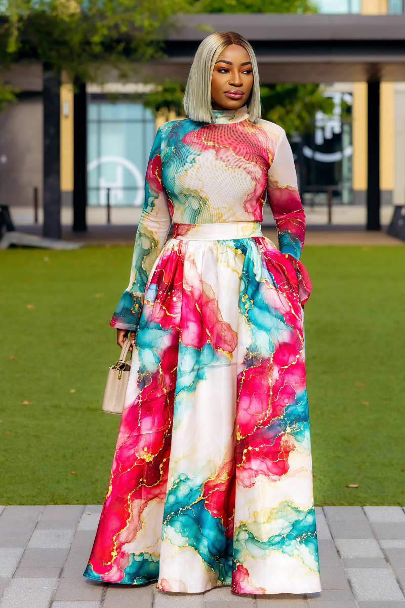 A woman stands outdoors on a paved walkway in front of a building and green lawn, wearing a long-sleeve maxi dress with a bold abstract watercolor print in pink, teal, blue, and cream. She is holding a small beige handbag.
