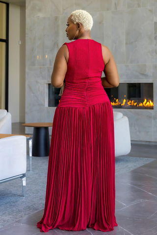 A woman with short blonde hair is shown from behind wearing a sleeveless, deep red pleated maxi dress in a modern indoor setting with a fireplace and light-colored stone walls.