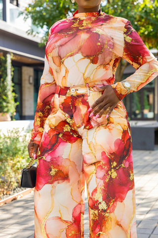 A woman wearing a long-sleeve high-neck jumpsuit with a bold abstract floral print in red, pink, orange, and gold on a cream base, photographed outdoors on a paved walkway.